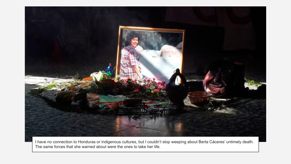 This image depicts a solemn and deeply symbolic memorial or altar, likely dedicated to Berta Cáceres, as suggested by the accompanying text. The overall scene is dark and somber, with a dramatic spotlight illuminating the